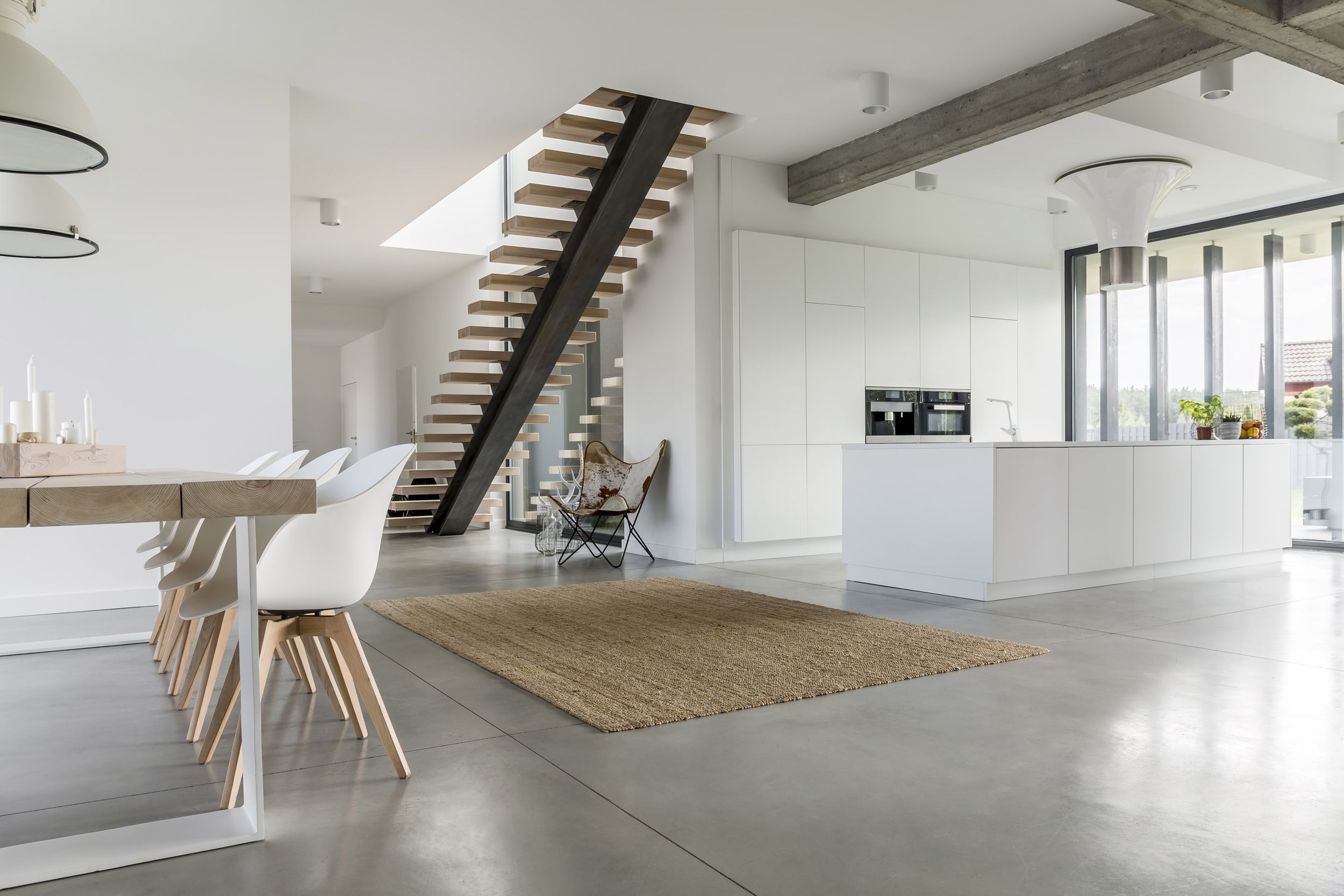 Offener moderner Wohnraum mit großem Holztisch, weißen Stühlen, Teppich, freitragender Treppe und Kücheninsel